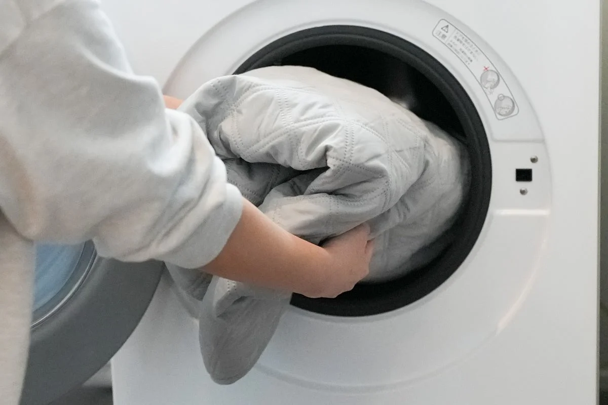 白いドラム式洗濯機に、キルティング加工された薄い色の大きな寝具を投入している様子