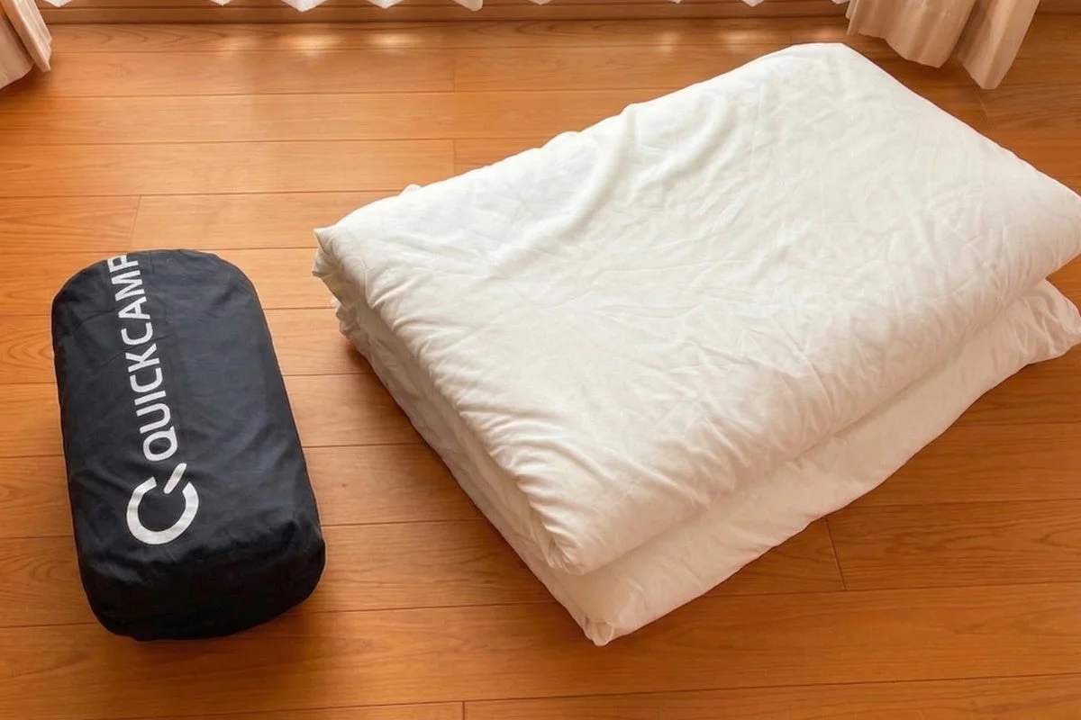 「QUICKCAMP」と書かれた黒い収納袋と、折りたたまれた白い布団が木の床に置かれています