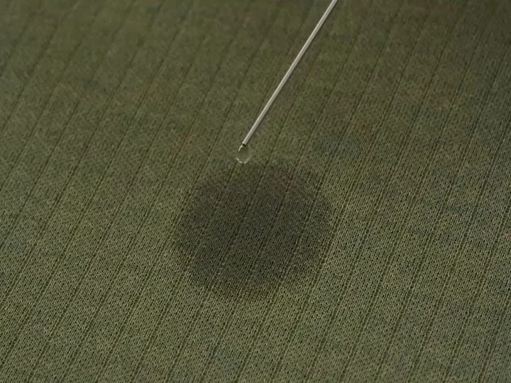 緑色の畝のある布地の上に、細い管から水滴が落ちようとしている瞬間を捉えた画像