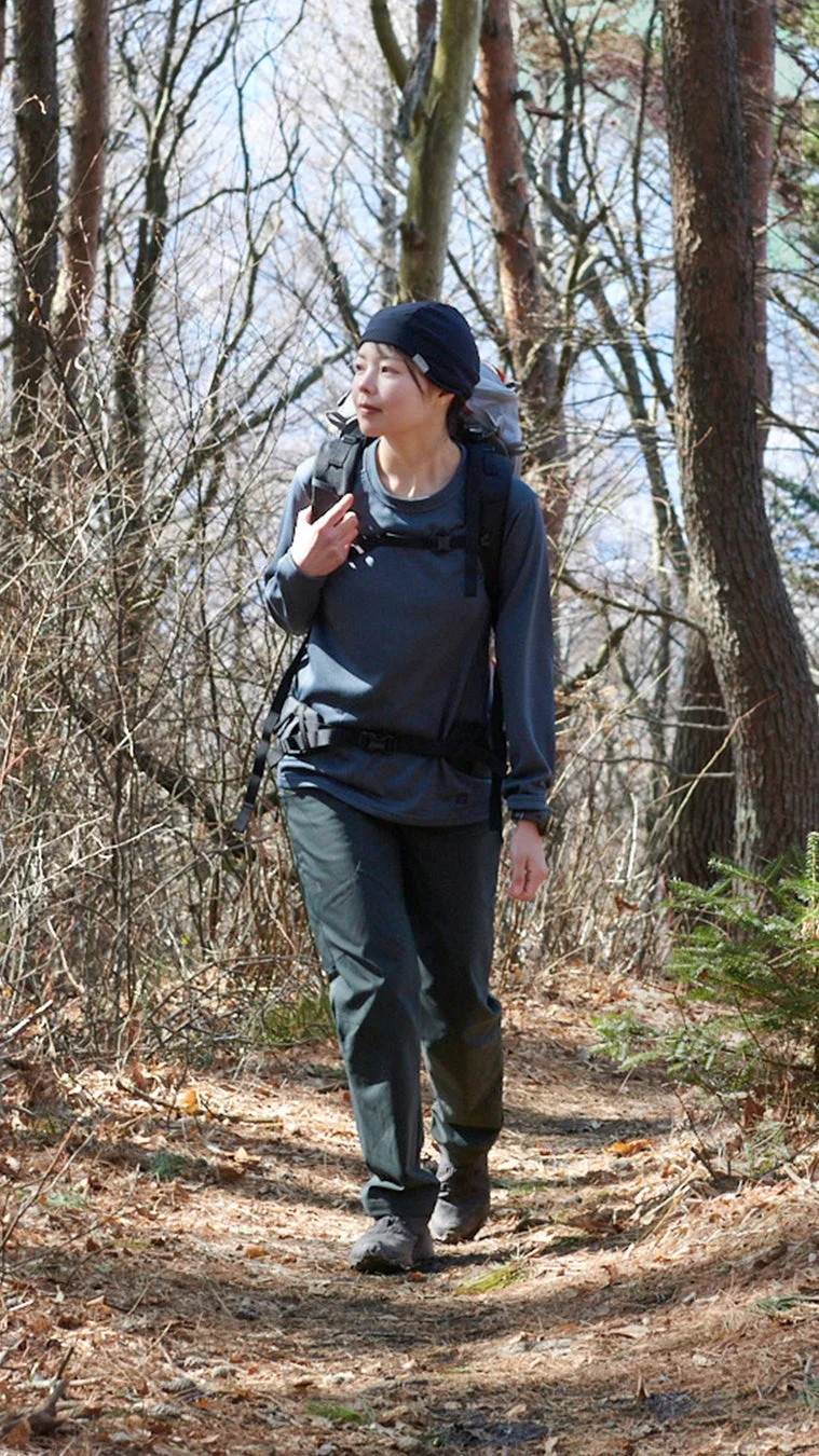 リュックサックを背負った女性が、冬から春にかけての裸の木々が立ち並ぶ森の中の小道を歩いています
