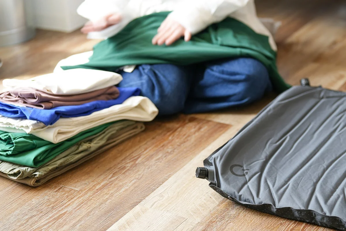 フローリングの床に座る人物の傍らに、カラフルな服の山とグレーのマットが置かれています