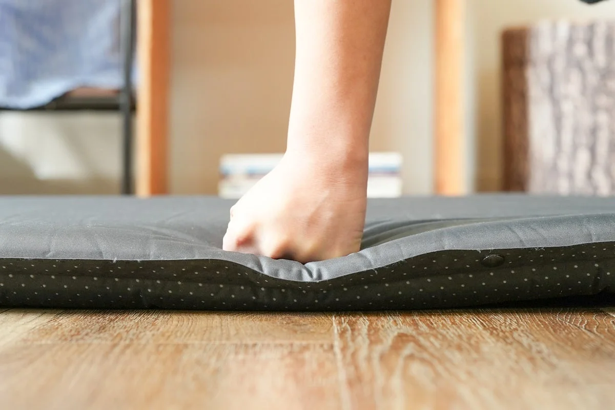 木目の床に置かれた黒いマットレスを人の手が押し、その柔らかさや弾力性を試している様子