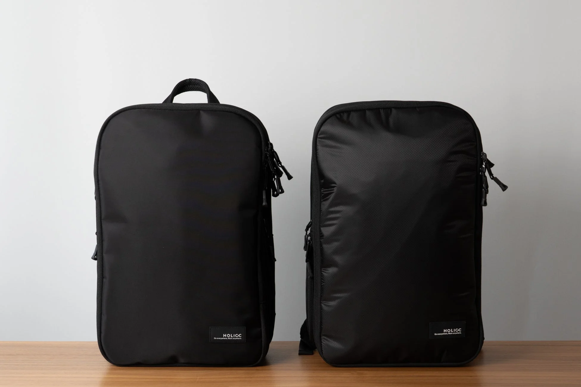 木製のテーブルの上に置かれた、黒いシンプルなデザインのバックパックが2つ写っています。
