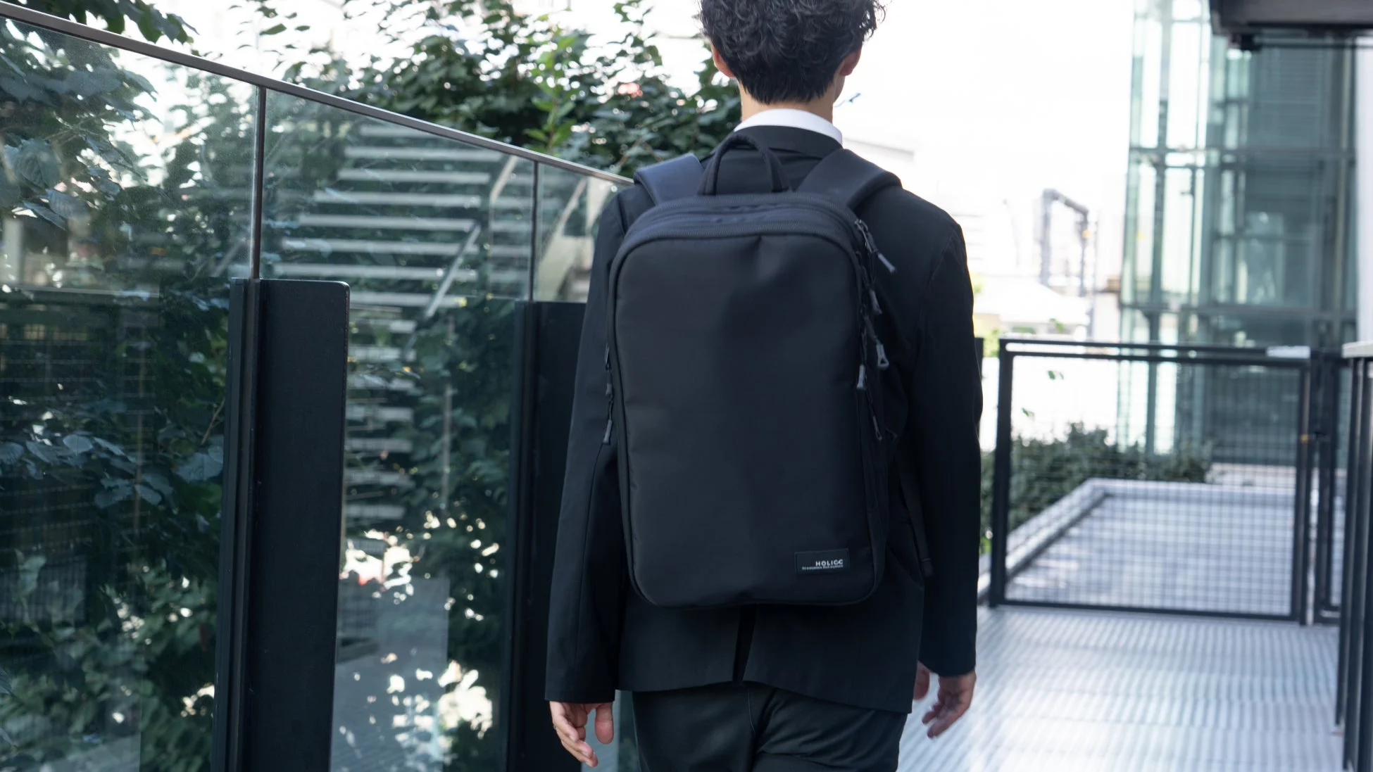 スーツ姿の男性が黒いバックパックを背負い、屋外の通路を歩いている後ろ姿です。